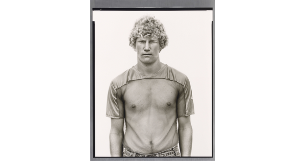A black-and-white photograph of a young White man with curly blonde hair wearing a see-through, mesh shirt.