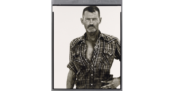 A black-and-white portrait photograph of a middle-aged White man with scruffy facial hair wearing a plaid button-down shirt standing with one hand on his hip.