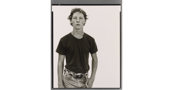 A black-and-white portrait photograph of a White teenage boy in a dark t-shirt and belted jeans with a large belt buckle, one hand hooked into his jeans pocket.