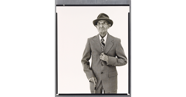 A black-and-white portrait photograph of an elderly White man in a hat, suit, tie, and one hand holding a cane.