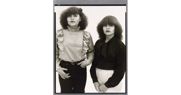 A black-and-white portrait photograph of two medium-skinned young women, both with teased dark hair and wearing ruffled blouses.
