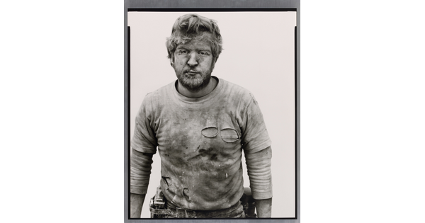 A black-and-white portrait photograph of a White man with messy hair and a beard wearing a dirty, ripped shirt.