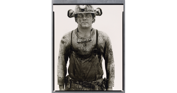 A black-and-white portrait photograph of a young White man covered in dirt wearing a hard hat and a shirt under coveralls.