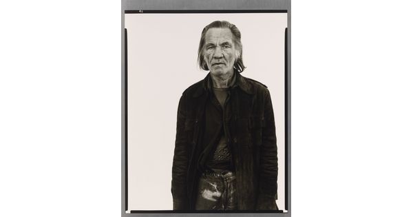 A black-and-white portrait photograph of an older White man with long hair wearing a partially-buttoned jacket over his clothing.