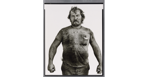 A black-and-white portrait photograph of a White man with messy hair and a beard wearing a dirty t-shirt and holding his arms out from his sides.