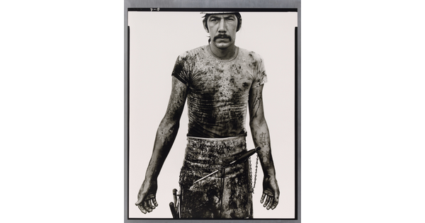 A black-and-white portrait photograph of a White man with a mustache wearing a dirty t-shirt and apron holding with different tools on it.