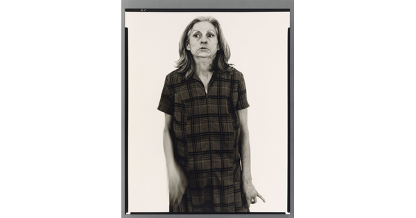 A black-and-white portrait photograph of an older White woman with medium-length gray hair, head held back slightly, wearing a house dress.