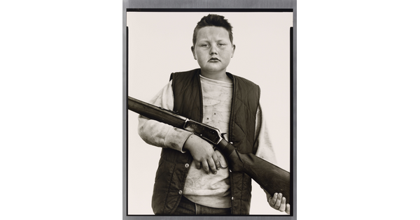 A black-and-white portrait photograph of a White boy with close cropped hair, wearing a quilted vest over a sweatshirt, and holding a rifle across his torso.