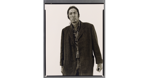 A black-and-white portrait photograph of a medium-skinned man with long hair pulled back, a goatee, and wearing a long wool coat.