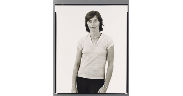 A black-and-white portrait photograph of a White woman with short hair, wearing a light V-neck shirt and dark pants.