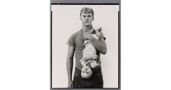 A black-and-white portrait photograph of a White man with light hair holding a baby upside down in front of his chest.