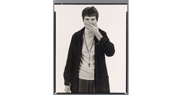 A black-and-white portrait photograph of a White woman with short, dark hair wearing a dark jacket and rosary beads around her neck holding one hand in front of her mouth and nose.