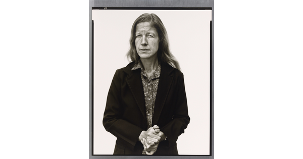 A black-and-white portrait photograph of a White woman with long hair wearing a button-down blouse under a suit jacket holding her hands clasped in front of her.