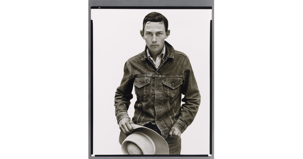 A black-and-white portrait photograph of a young White man with short dark hair wearing a denim jacket and holding a cowboy hat in front of him.