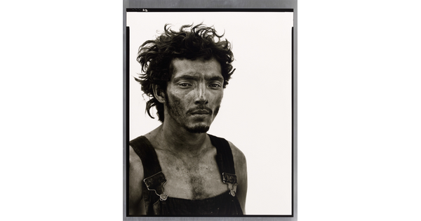 A black-and-white portrait photograph of a man from the chest up with messy hair, a dirty face, and overalls.