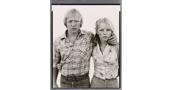 A black-and-white portrait photograph of a White teenage boy wearing a plaid button-down shirt, his arm around the shoulders of a White teenage girl wearing a sleeveless shirt.