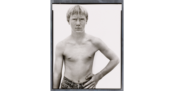 A black-and-white photograph of a shirtless White boy with light hair and a freckled face standing with one hand on his hip.