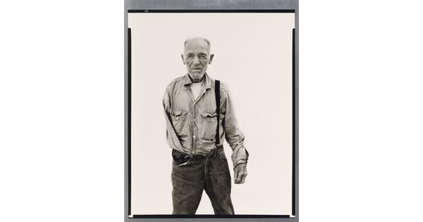 A black-and-white portrait photograph of an older, balding White man with one arm wearing a button-down shirt and jeans held up with one suspender.