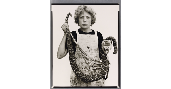 A black-and-white portrait photograph of a White teenage boy with curly hair wearing a dirty apron and holding a gutted snake in front of him.