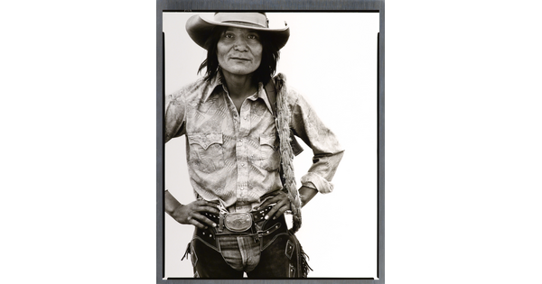 A black-and-white portrait photograph of a Native American man with long hair wearing a cowboy hat, button-down shirt, jeans, and chaps, standing with hands on hips.