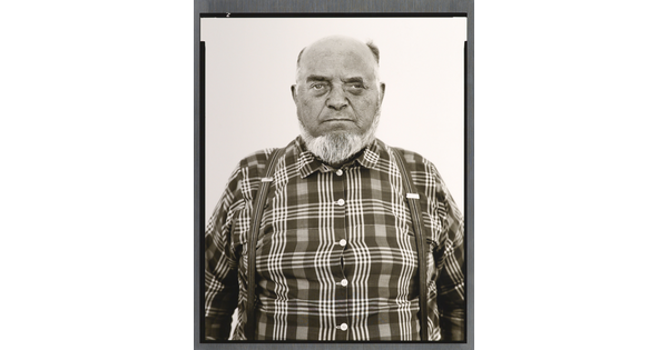 A black-and-white portrait photograph of an older, balding White man with a gray beard wearing a plaid button-down shirt and suspenders.