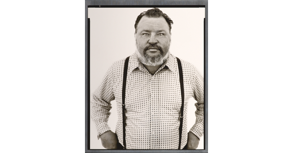 A black-and-white portrait photograph of a middle-aged White man with a receding hairline and salt-and-pepper beard wearing a checked button-down shirt and suspenders.