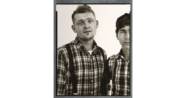 A black-and-white portrait photograph of two White men, both in similar plaid shirts and suspenders, standing shoulder to shoulder.