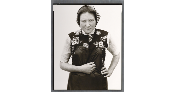 A black-and-white portrait photograph of young White woman wearing a checked kerchief, flowered vest, and dark apron.