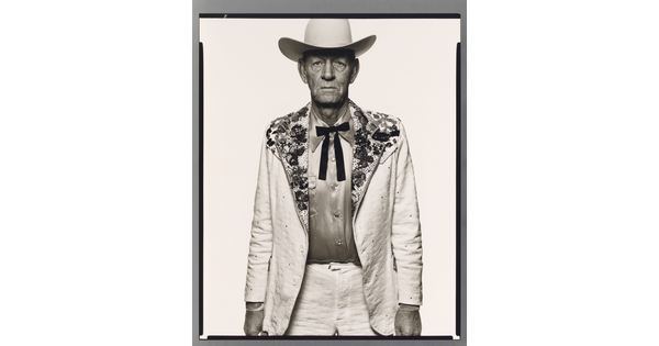 A black-and-white portrait photograph of an older White man wearing a white cowboy hat, a white suit jacket with rhinestones on the collar, a satin shirt, and a bow tie.
