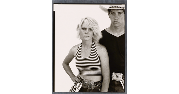 A black-and-white portrait photograph of a young White woman wearing a striped halter top, one hand on her hip, and a young White man wearing a cowboy hat and V-neck t-shirt, both wearing large buckles on their belts.