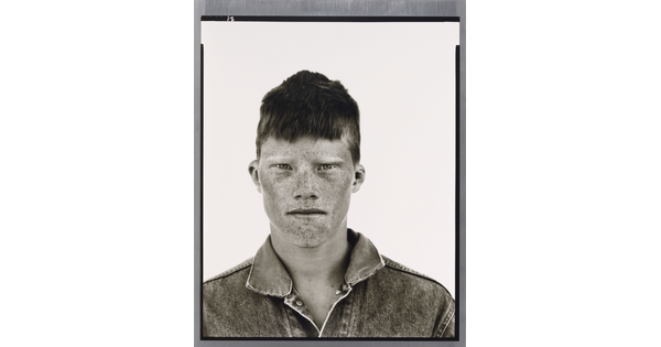 A black-and-white portrait photograph of a White teenage boy, face covered in freckles, short cropped hair, wearing a denim jacket.