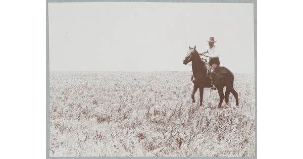 A black-and-white photograph of a man wearing a cowboy hat riding a horse in an open field.