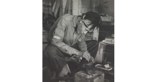 A black-and-white photograph of a seated Native American man bent over an anvil in a workshop.