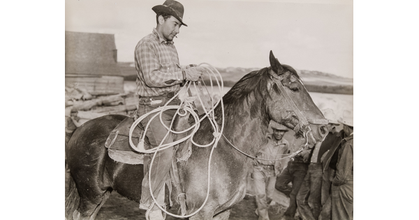 A black-and-white photograph of a light-skinned man in a brimmed hat holding a lasso on horseback.
