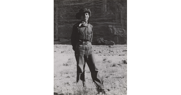 A black-and-white photograph of a man wearing a cowboy hat and neckerchief posing in front of what looks like the face of a cliff.