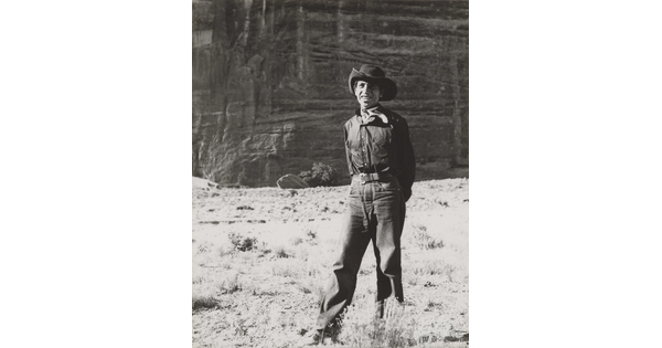 A black-and-white photograph of a man wearing a cowboy hat and neckerchief posing in front of what looks like the face of a cliff.