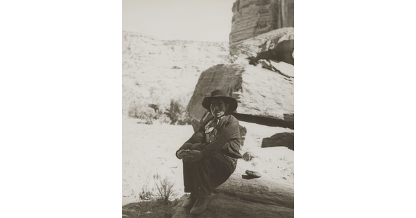 A black-and-white photograph of a man wearing a cowboy hat and neckerchief sitting on a rock in the shadows of a cliff face.
