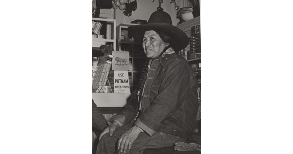 A black-and-white photograph of a Native American person wearing a jean jacket and cowboy hat sitting on a counter in a general store.