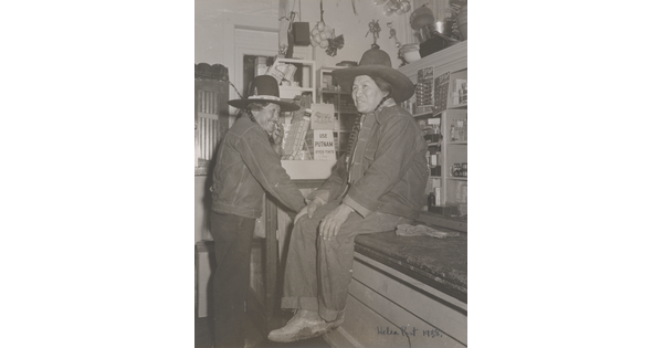 A black-and-white photograph of two Native American people in a general store: one wearing a jean jacket and cowboy hat sitting on a counter and the other dressed similarly standing nearby.
