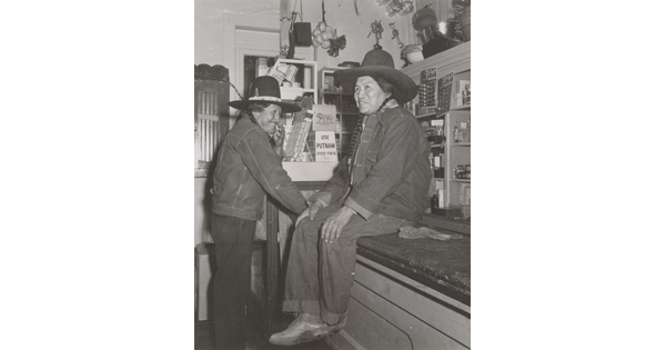 A black-and-white photograph of two Native American people in a general store: one wearing a jean jacket and cowboy hat sitting on a counter and the other dressed similarly standing nearby.