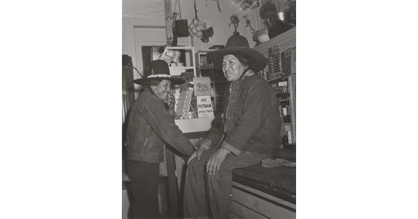 A black-and-white photograph of two Native American people in a general store: one wearing a jean jacket and cowboy hat sitting on a counter and the other dressed similarly standing nearby.