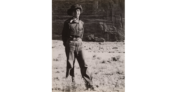 A black-and-white photograph of a man wearing a cowboy hat and neckerchief posing in front of what looks like the face of a cliff.