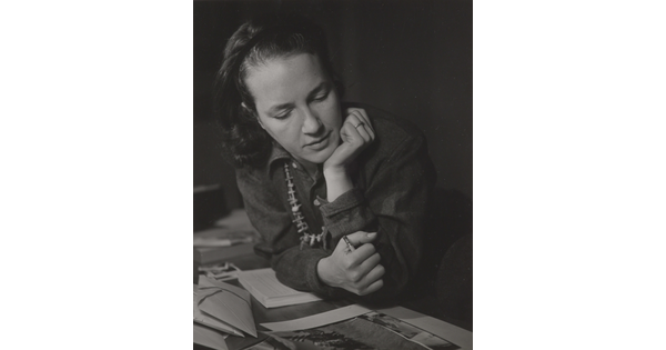 A black-and-white photograph of a White woman with her hand under her chin looking down at a photograph. 
