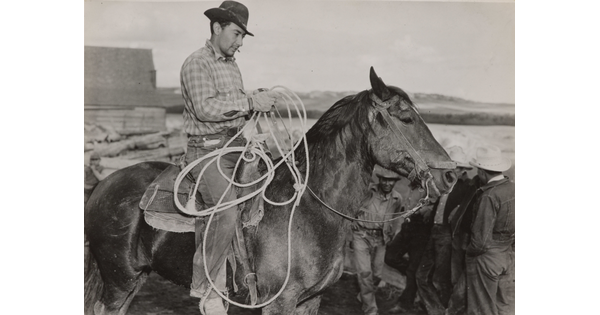 A black-and-white photograph of a light-skinned man in a brimmed hat holding a lasso on horseback.