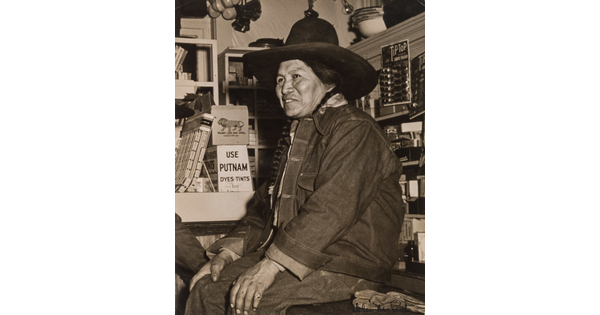 A black-and-white photograph of a Native American person wearing a jean jacket and cowboy hat sitting on a counter in a general store.
