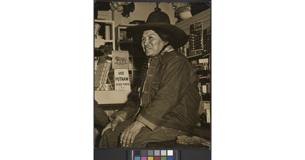 A black-and-white photograph of a Native American person wearing a jean jacket and cowboy hat sitting on a counter in a general store.