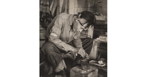 A black-and-white photograph of a seated Native American man bent over an anvil in a workshop.