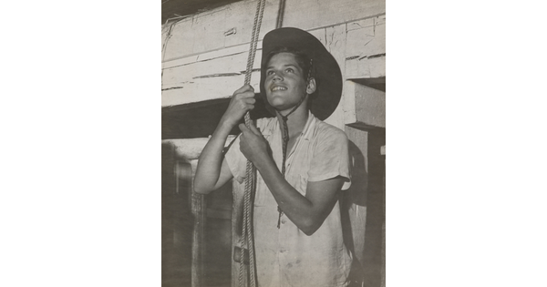 A black-and-white photograph of a medium-skinned boy in a cowboy hat holding a couple of ropes suspended from above.