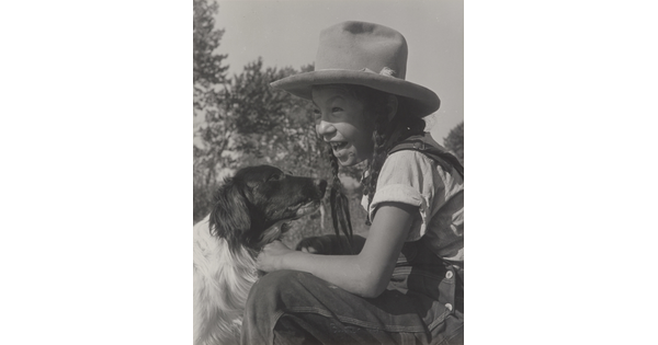 A black-and-white photograph of a smiling Native American child wearing a Homburg hat and sitting in front of a black-and-white dog.