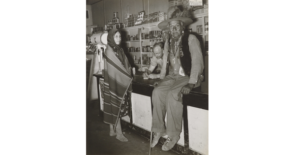 A black-and-white photograph of a Indigenous man sitting on a counter, an Indigenous woman wrapped in a blanket standing at the counter, and a White man behind the counter.